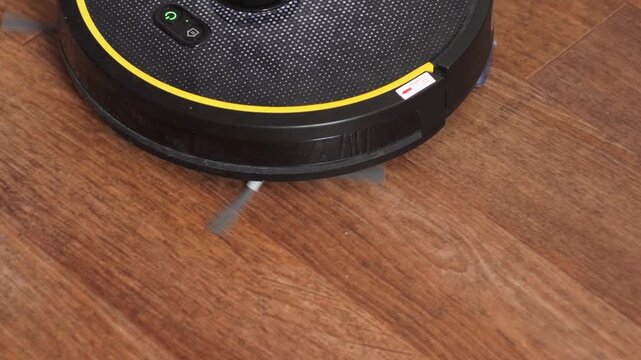 Black robotic vacuum cleaner cleaning wooden laminate floor in modern apartment. Smart home gadget for automated domestic housework and floor care