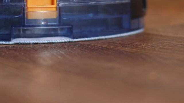 Robotic vacuum cleaner performing wet cleaning on laminate flooring in a modern apartment. Close up of automatic cleaner brushes and water tank module in motion
