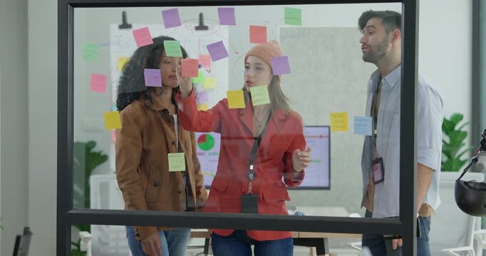 Diverse group of startup team engaging in brainstorming session, using sticky notes on glass wall to organize ideas, plan next steps. Creativity and collaborative spirit of modern entrepreneurial team