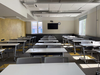 Desks and chairs are set in rows inside a classroom. There is a clock on the wall. Sunlight enters through the windows. The room is empty during the day