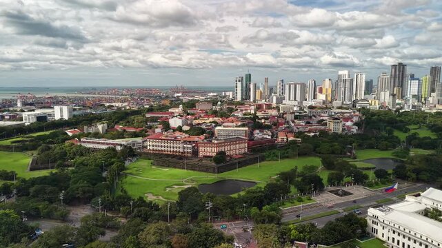 Wide aerial panorama captures Intramuros walled city with stone fortifications and green moat spaces showing colonial compound with red rooftops and modern Manila skyline under dramatic sky