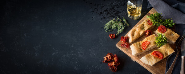 Freshly baked Italian focaccia bread pieces with tomatoes and fresh herbs on black countertop....