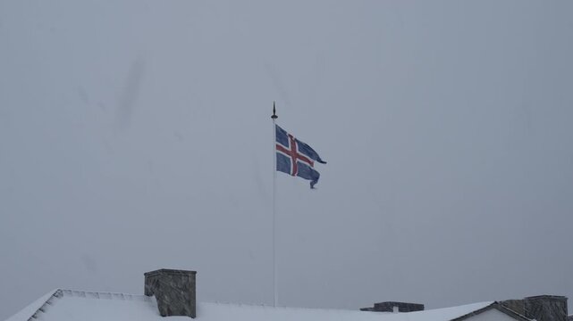 Icelandic flag waving in the wind over a snowy rooftop during a blizzard.