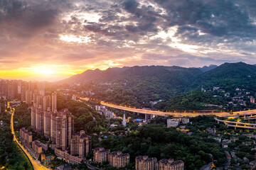 Aerial view of high-rise residential buildings and highway overpass nestled in lush green mountains at sunset in Chongqing, China.