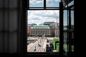 View through an open dark window of a sunny city bridge lined with flags leading to grand historic buildings in Stockholm Sweden © Michael Persson