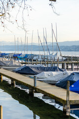 Fototapeta premium Vertical view of a wooden pier with moored sailboats on Lake Zurich (Zürichsee) in Switzerland, featuring the distant snow-capped Swiss Alps under a clear sky.