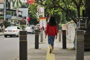 Young Woman in Red Sweater Walking on Jakarta Sidewalk, Urban Pedestrian Scene with Tactile Paving for Blind, City Life in Indonesia