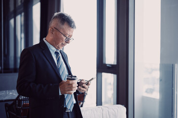Portrait of an Asian senior businessman holding coffee paper cup and texting on a smartphone while standing near a big window.
