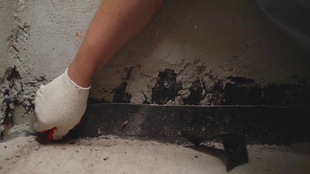 Close-up of a worker's hands in gloves applying waterproofing mastic to a concrete wall base with a trowel. Renovation, repair, and construction concept. Sealing a joint.