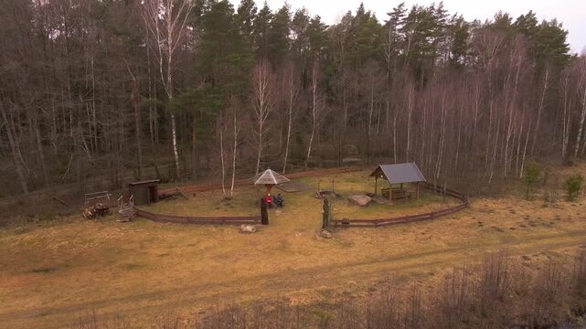 Aerial view shows a fenced lakeside picnic clearing in Belarus with a canopy, pavilion, firepit, woodpile, and dock. People sit and walk as the camera pulls back.