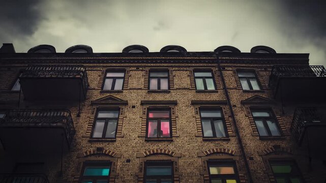 Eerie facade of an old brick apartment building at dusk, featuring windows illuminated with dramatically colored light and silhouetted figures.