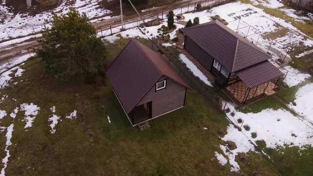 Aerial drone passes over a Belarusian dacha with two wooden cottages, brown metal roofs, pine, greenhouse, fenced yard, muddy lane, and power lines in late winter