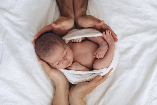 Newborn baby wrapped in soft cloth, cradled gently by adult hands on white bedding