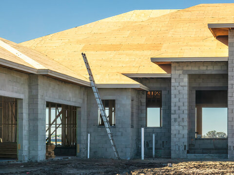 Part of a house and garage under construction, with concrete shell in morning shadow and an extension ladder leaning against a pitched roof covered with plywood, in a suburban development in Florida