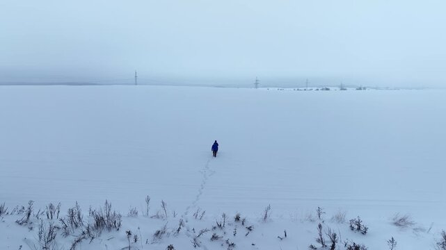Aerial view of a snow plain in Belarus as two figures walk away, leaving one trail. Electrical pylons line the horizon, birch trees sit left, shrubs dot the field in fog.