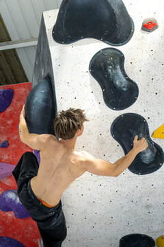 young muscular white male indoor climbing on bouldering wall with no top and black jogger baggy pants on overhang wall looking up to next big dyno move and left heel hook in with brown hair