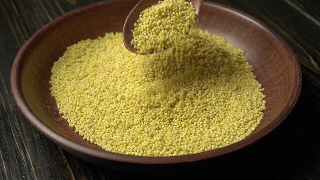Spoonful of yellow millet grains being poured into a wooden bowl, showcasing the texture and color of the seeds in a rustic kitchen setting