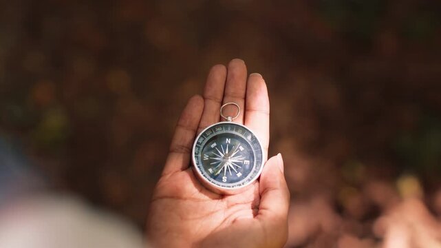 A hand holds a compass while standing on the forest floor. The background shows blurred trees and leaves, indicating an outdoor exploration activity.