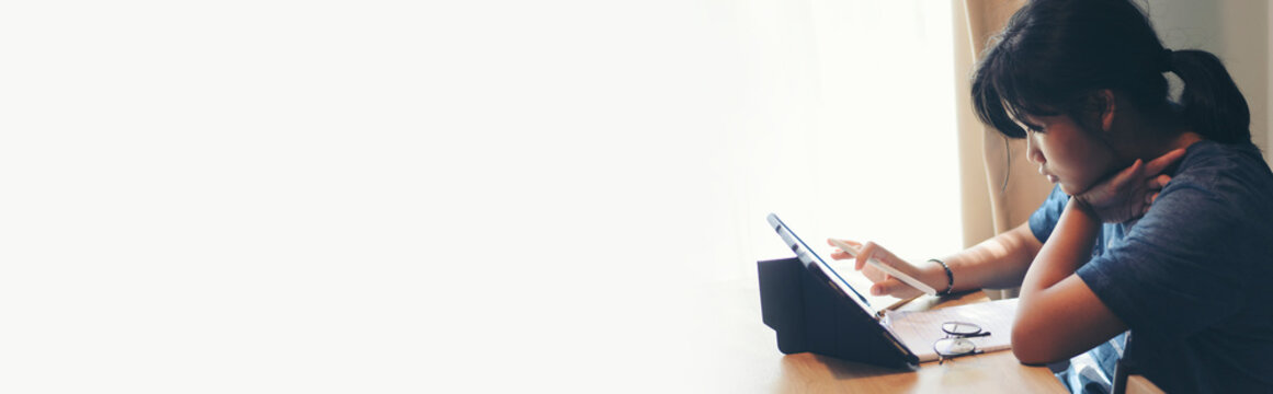 Young Asian student using tablet for online learning at home. Girl studying digital lesson with white copy space for banner. Concept of e-learning and modern education.