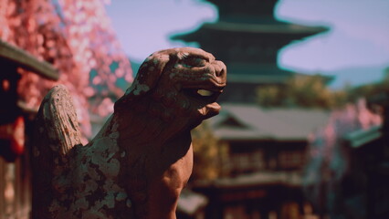 A weathered guardian statue stands proud amidst blooming cherry trees, offering a glimpse of traditional Japanese architecture in the background, creating a serene atmosphere in spring. © icetray