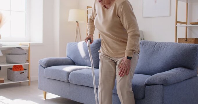 Elderly senior woman with cane, hip pain at home. She sits on sofa in a bright room, gripping her hip, grimacing, needing a crutch or cane. Clear concept pain, injury, limited mobility in retirement.