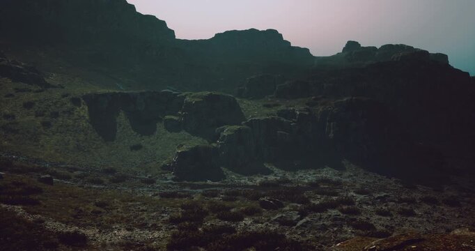 A vast mountain terrain is seen covered in grayish hues, with rocky formations towering over a sparsely vegetated area during the twilight hours, creating a mysterious atmosphere.