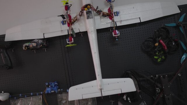 White model airplane with multiple drone propellers and components attached, resting on a dark surface alongside remote control accessories and additional drone parts