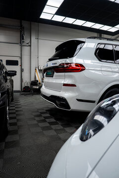 White BMW X7 xDrive30d rear three quarter in a detailing bay, LED tail lamps, chrome dual exhausts, roof rails, and dark rear glass, framed by two cars and grid ceiling
