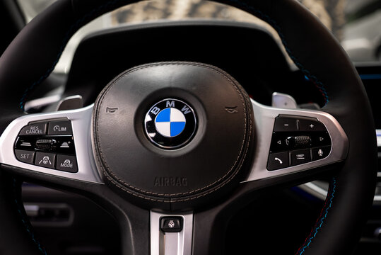 Close view of BMW X7 steering wheel shows roundel on stitched leather, metal accents, tri color stitching, cruise and drive toggles, audio and phone controls, heated button under soft light.