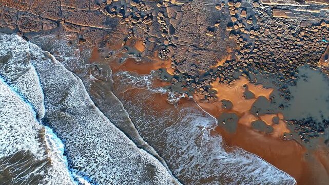 Top down aerial shot of rocky shoreline and ocean waves. Vertical view of limestone rock formations and sandy beach with incoming waves at Southerndown coast.