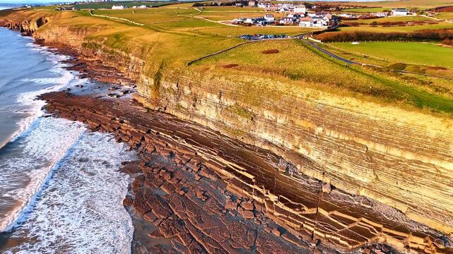 Coastal landscape of green fields and limestone cliffs in Wales. Aerial perspective of Southerndown cliffs with village buildings and green meadows overlooking the sea.
