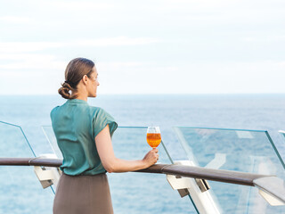 Stylish woman with a glass of drink standing on the empty deck of a cruise ship against the backdrop of sea waves. Sunny day. Chill lifestyle, luxury travel. Perfect for holiday and travel themes © Svetlana