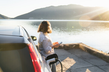 Fototapeta premium Woman traveler with charging plug next to her electric car on the sea coast at sunset