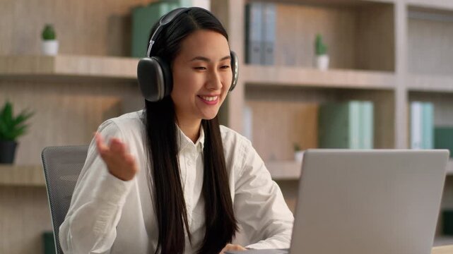 Asian business woman office worker talking video call headphones laptop greeting smiling Japanese Chinese girl ethnic female businesswoman Korean operator in headset online webcam use computer indoors