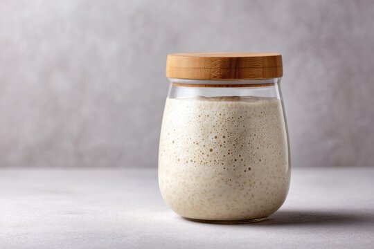Active sourdough starter in a clear glass jar with a wooden lid placed on a light gray surface against a soft textured background