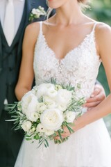 Bride in elegant white lace wedding dress holds bouquet of white flowers while standing close to groom in a black suit, lush greenery visible in the background