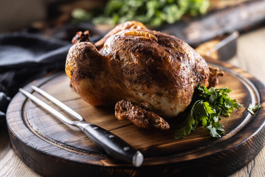 A whole chicken, baked until golden brown, is served on a wooden cutting board, garnished with fresh parsley and accompanied by a meat fork in a rustic setting