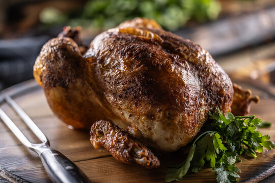 A whole chicken, baked until golden brown, is served on a wooden cutting board, garnished with fresh parsley and accompanied by a meat fork in a rustic setting