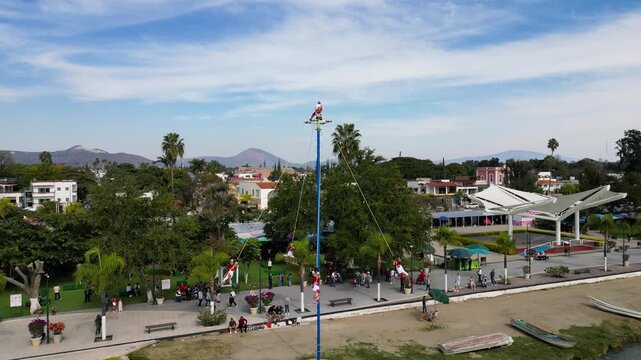 Aerial view of the ancient Papantla Flyers ritual in Chapala, Mexico. Performers descend from a tall pole above spectators at a vibrant lakeside promenade