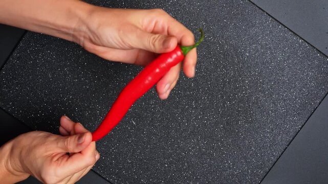 Hands holding whole red hot chili pepper over board. Overhead shot of person displaying a long red spicy pepper over a black speckled cutting board.