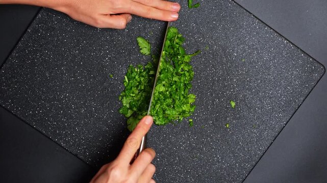 Hands finely chopping fresh green cilantro with chef knife. Top view of person mincing coriander herbs on a dark textured cutting board using a professional blade.