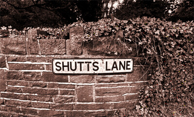 A sepia toned street sign for Shutts Lane rests against an old stone wall, half‑claimed by trailing ivy. The warm vintage palette deepens the sense of age and quiet charm in Norwood Green, Halifax. UK