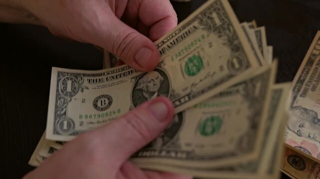 Close-up of an elderly man slowly counting one dollar bills at a table. His aged hands carefully separate small denomination banknotes, reflecting modest pension income, financial caution and everyday