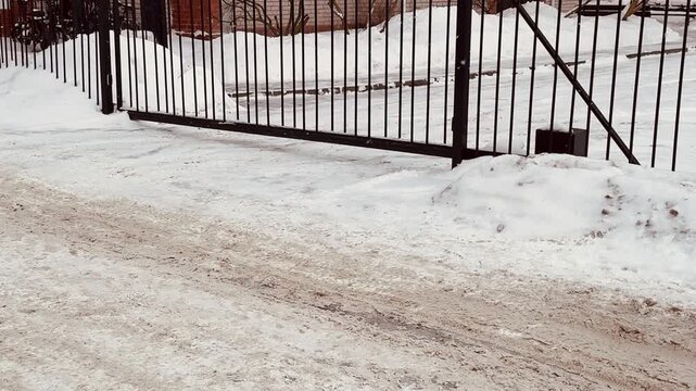 Snow covered iron gate slides open, suburban brick house entrance, automatic motor visible, keypad on post, frosty driveway.