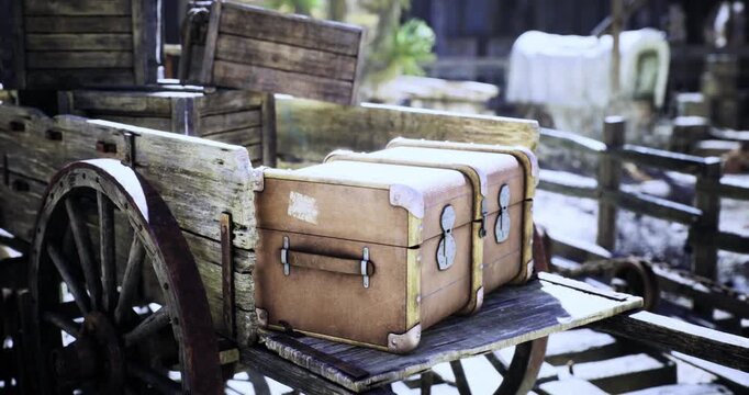Two leather suitcases sit on a weathered wooden cart, surrounded by rustic western elements. The background features a blurred outline of a ghost town ambiance, emphasizing nostalgia.