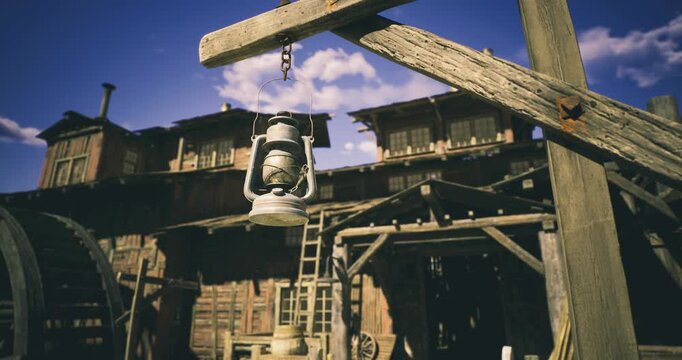 A vintage lantern is suspended from a wooden post in a historic outdoor area. In the background, a weathered wooden structure with multiple windows and a ladder is visible under a clear blue sky.