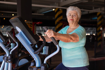 Fototapeta premium Active senior woman exercising on elliptical trainer in gym. Concept of menopause fitness, bone health, osteoporosis prevention and healthy aging. Mature woman maintaining strength and mobility 