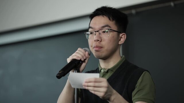 A young Asian male wearing glasses speaks into a handheld microphone while referencing cue cards, ideal for public speaking, student presentation, or speech preparation concepts.