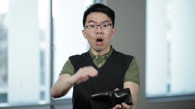 A young Asian male wearing glasses scratches his head with a worried expression while looking at an empty wallet, ideal for financial stress, budgeting, or money problems concepts.