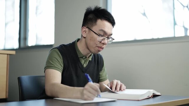 A young Asian male student wearing glasses writes in a notebook while studying from a textbook at a desk, ideal for studying, homework, or academic focus concepts.